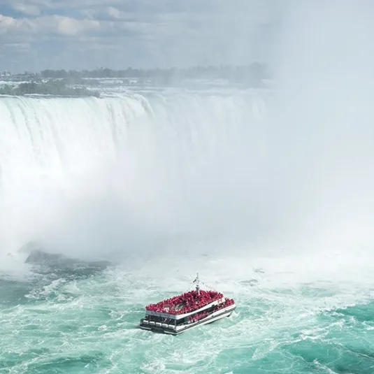 A tour boat filled with people in red ponchos approaches the misty base of Niagara Falls against a backdrop of powerful cascading water.