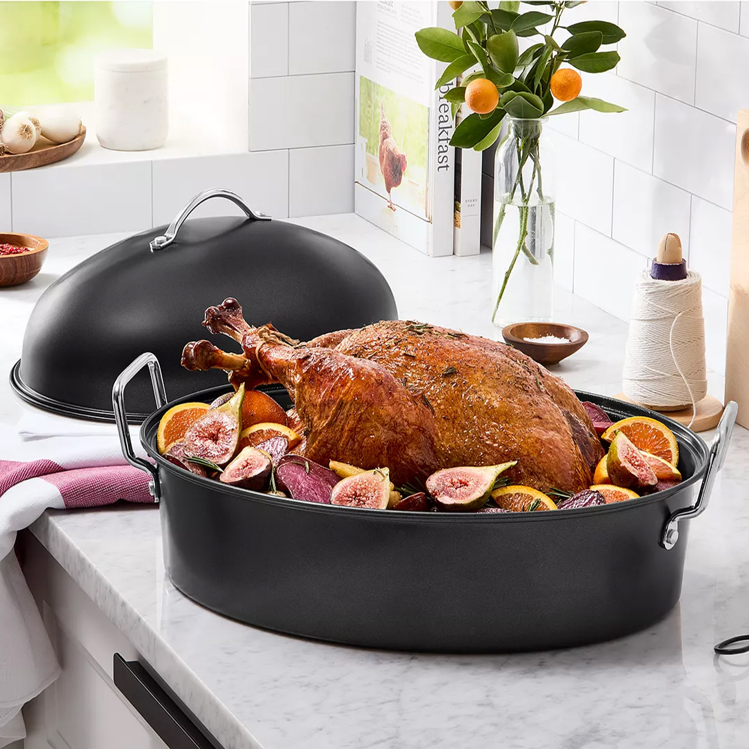 A dark roasting pan with a lid, holding a cooked turkey surrounded by figs and citrus slices, sits on a kitchen counter. Nearby are spices, kitchen twine, and citrus plant decor.