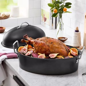 A dark roasting pan with a lid, holding a cooked turkey surrounded by figs and citrus slices, sits on a kitchen counter. Nearby are spices, kitchen twine, and citrus plant decor.