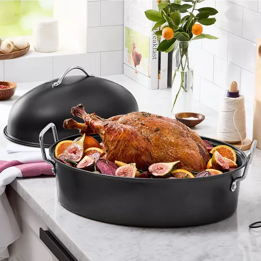 A dark roasting pan with a lid, holding a cooked turkey surrounded by figs and citrus slices, sits on a kitchen counter. Nearby are spices, kitchen twine, and citrus plant decor.