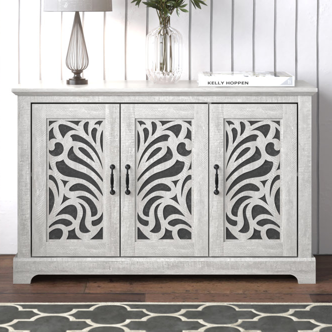 A vintage-inspired sideboard buffet with ornate, floral scrollwork patterns on the cabinet doors and a light gray finish. It has four metal handles and a simple tabletop with a lamp and a book.