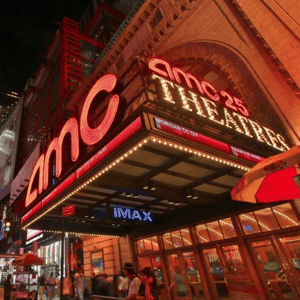 AMC 25 Theatres marquee at night with bright neon lights, prominently featuring IMAX. Pedestrians and nearby street vendors are visible on the bustling street.