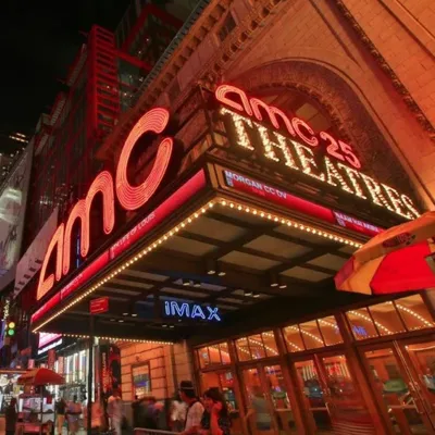 AMC 25 Theatres marquee at night with bright neon lights, prominently featuring IMAX. Pedestrians and nearby street vendors are visible on the bustling street.