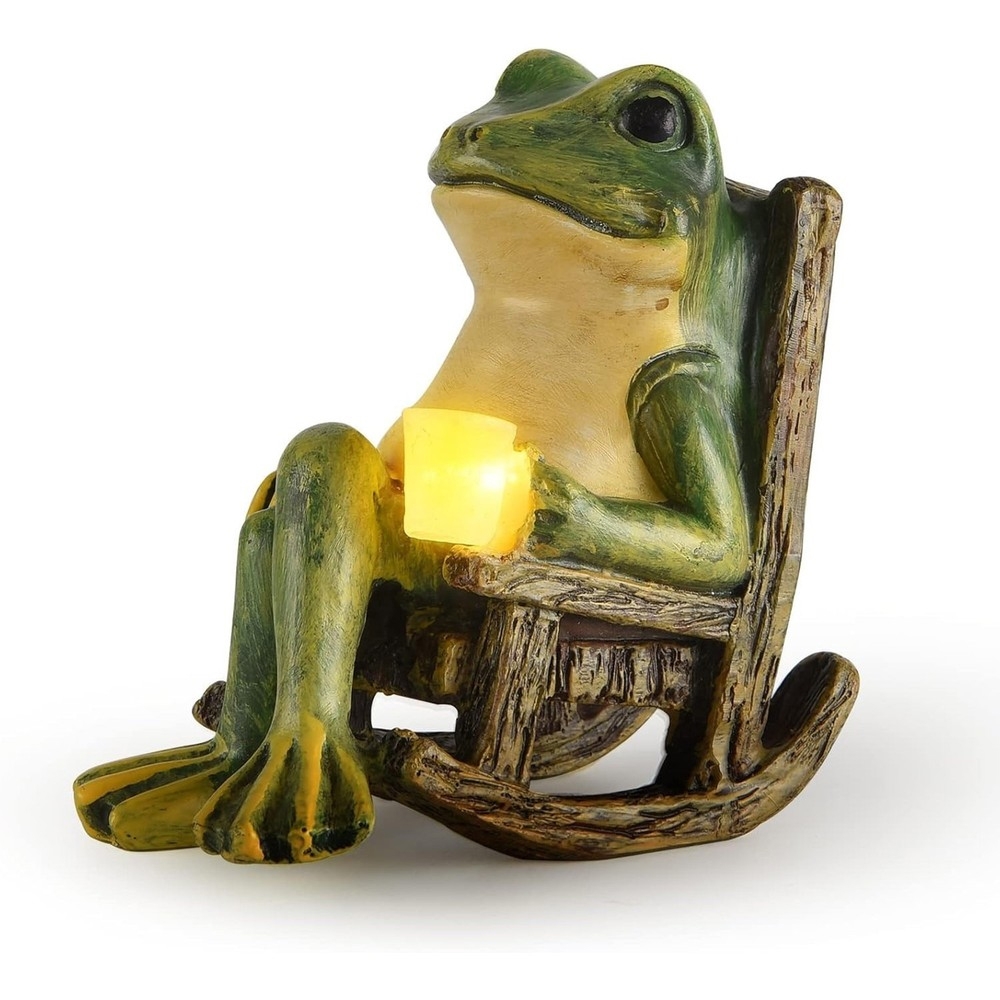 A whimsical frog statue sitting in a rocking chair holds a glowing yellow light, designed for use as a solar-powered garden decoration.