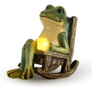 A whimsical frog statue sitting in a rocking chair holds a glowing yellow light, designed for use as a solar-powered garden decoration.