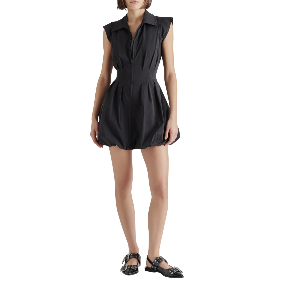 A woman wears a black sleeveless, collared dress with a zip front and a bubble hem, paired with black flats featuring metal buckle details.