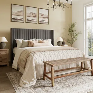 A cozy bedroom featuring a dark upholstered bed with a beige quilt and throw pillows, framed landscape art above, wooden nightstands with lamps, a wicker bench at the foot, a patterned rug, and a potted plant near a large window.