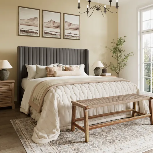 A cozy bedroom featuring a dark upholstered bed with a beige quilt and throw pillows, framed landscape art above, wooden nightstands with lamps, a wicker bench at the foot, a patterned rug, and a potted plant near a large window.