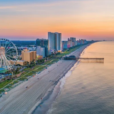 A beachside cityscape featuring a ferris wheel, high-rise buildings, a pier, and a coastline at sunset.