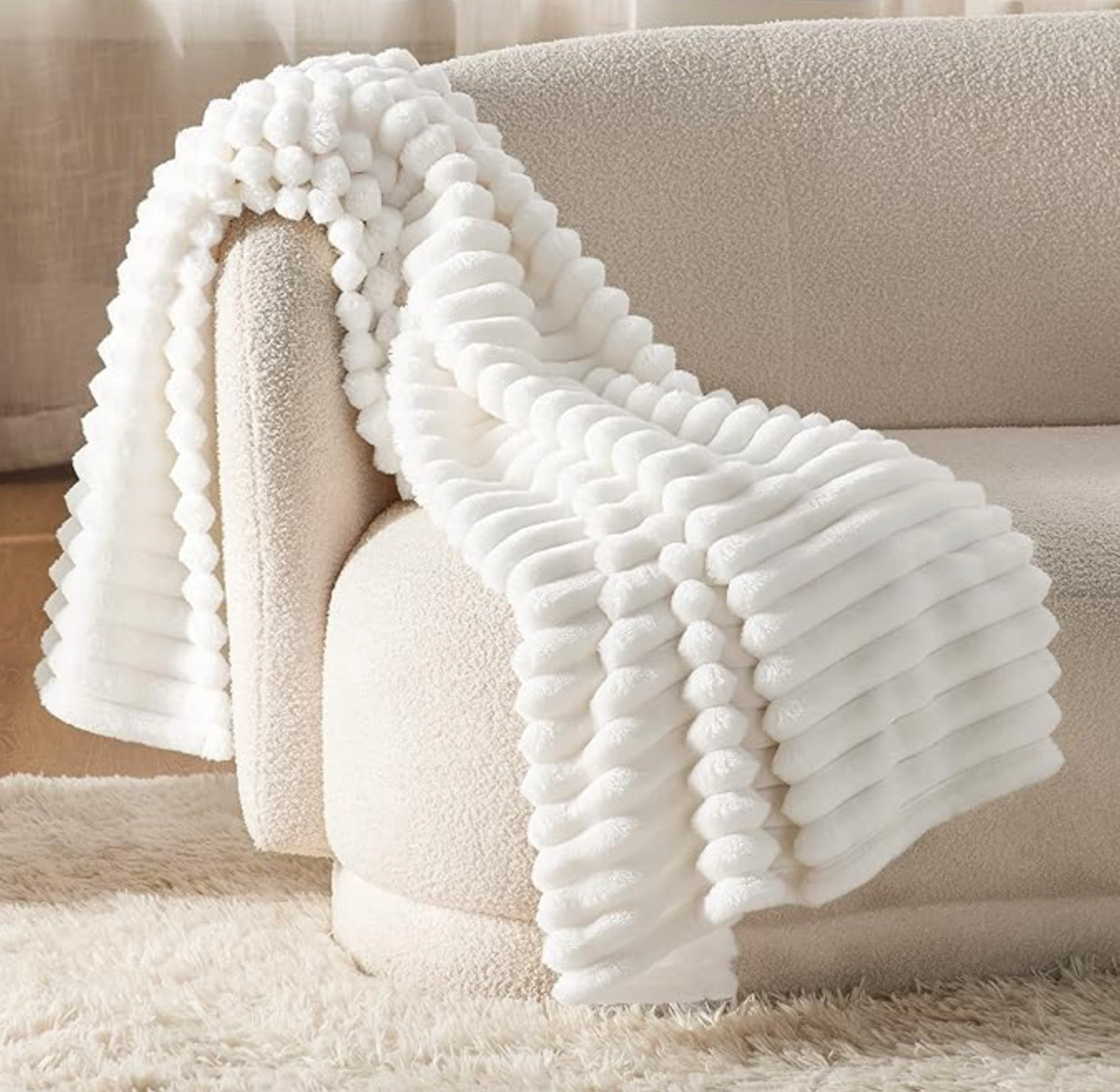 A white textured fleece blanket with a ribbed, ridged design is draped over a cream sofa, surrounded by a plush rug.