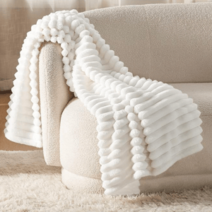 A white textured fleece blanket with a ribbed, ridged design is draped over a cream sofa, surrounded by a plush rug.