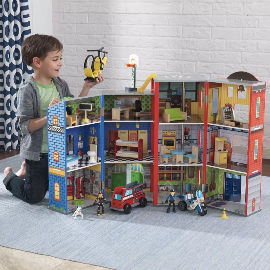 A child is playing with a multi-story toy fire station set, featuring detailed rooms, a helicopter, fire engine, motorcycle, and firefighter figures.