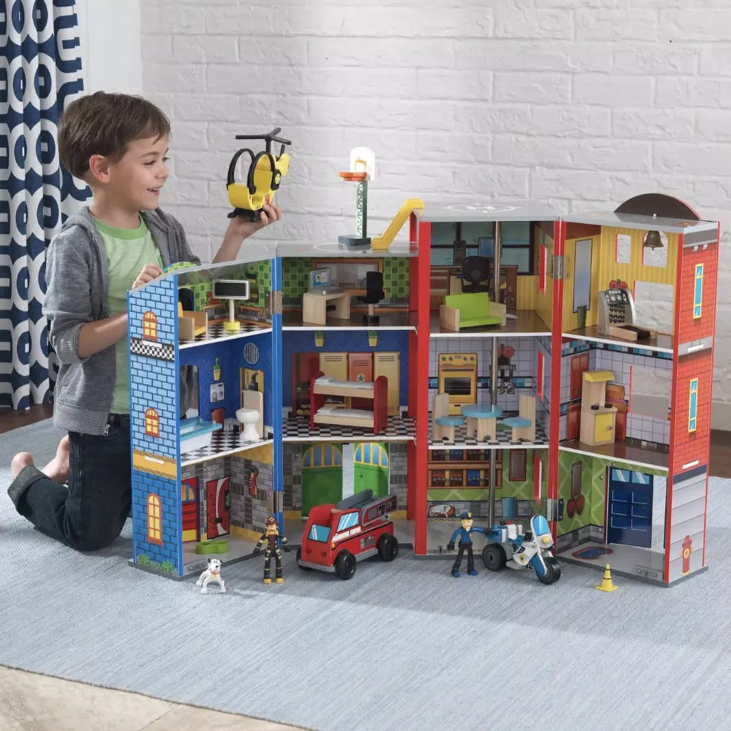 A child is playing with a multi-story toy fire station set, featuring detailed rooms, a helicopter, fire engine, motorcycle, and firefighter figures.