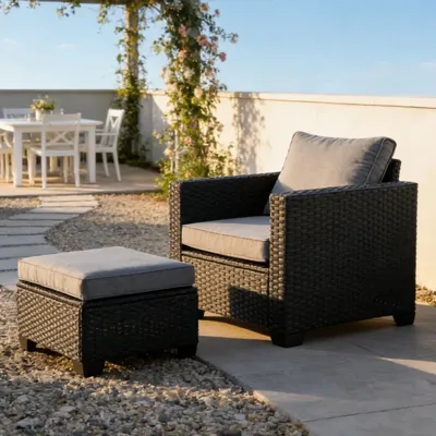 A patio setup featuring a dark wicker armchair with gray cushions and a matching ottoman, placed on a gravel surface. In the background, there's a dining table under a pergola adorned with climbing flowers.