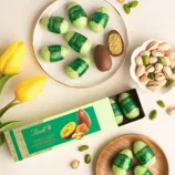 Green-wrapped chocolates labeled as Dubai Style Chocolate with pistachio fillings are displayed alongside a few whole pistachios and yellow tulips.