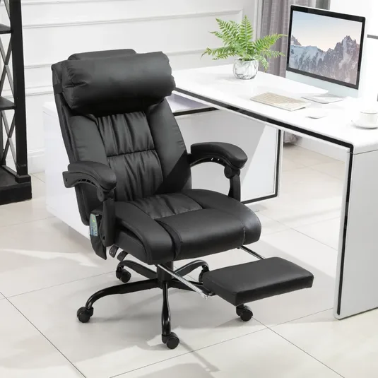 A black ergonomic office chair with an adjustable headrest and a retractable footrest is placed in front of a white desk with a computer monitor, keyboard, and a potted plant on top.