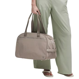 A person carries a taupe-colored duffel bag with two handles, wearing light green pants and sandals.