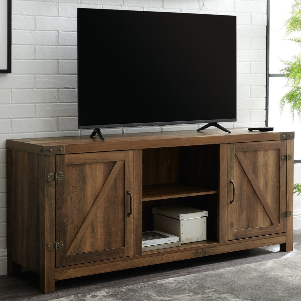 A wooden TV stand with a rustic design features two side cabinets with metal hinges and handles, and a central open shelf. A flatscreen TV sits on top, placed against a white brick wall. A shelf above holds small decor items and framed pictures.