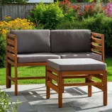 A wooden patio furniture set consisting of a corner bench with gray cushions and a matching ottoman, surrounded by a garden with flowers and greenery.