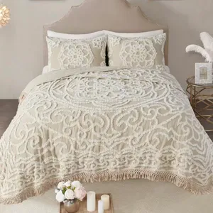 A bedding set featuring a textured, ornate cream pattern with a fringed edge, accompanied by two matching pillow shams. A small round side table holds a decorative vase with feathers and a photo frame, while a tray with flowers and candles is placed at the foot of the bed.