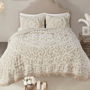 A bedding set featuring a textured, ornate cream pattern with a fringed edge, accompanied by two matching pillow shams. A small round side table holds a decorative vase with feathers and a photo frame, while a tray with flowers and candles is placed at the foot of the bed.