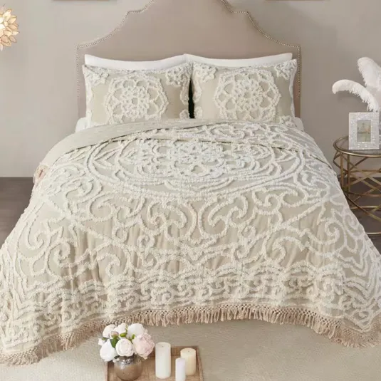 A bedding set featuring a textured, ornate cream pattern with a fringed edge, accompanied by two matching pillow shams. A small round side table holds a decorative vase with feathers and a photo frame, while a tray with flowers and candles is placed at the foot of the bed.