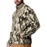 A man is wearing a khaki and brown camouflage-patterned Columbia fleece jacket with a high collar. The jacket has a patch logo on the chest and is paired with brown pants.