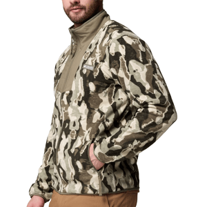 A man is wearing a khaki and brown camouflage-patterned Columbia fleece jacket with a high collar. The jacket has a patch logo on the chest and is paired with brown pants.