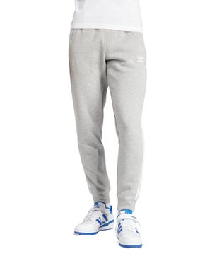 Gray Adidas Men's Adicolor Pants with a slim fit, featuring the Adidas logo on the upper thigh and white stripes along the sides. Paired with white sneakers with blue accents.