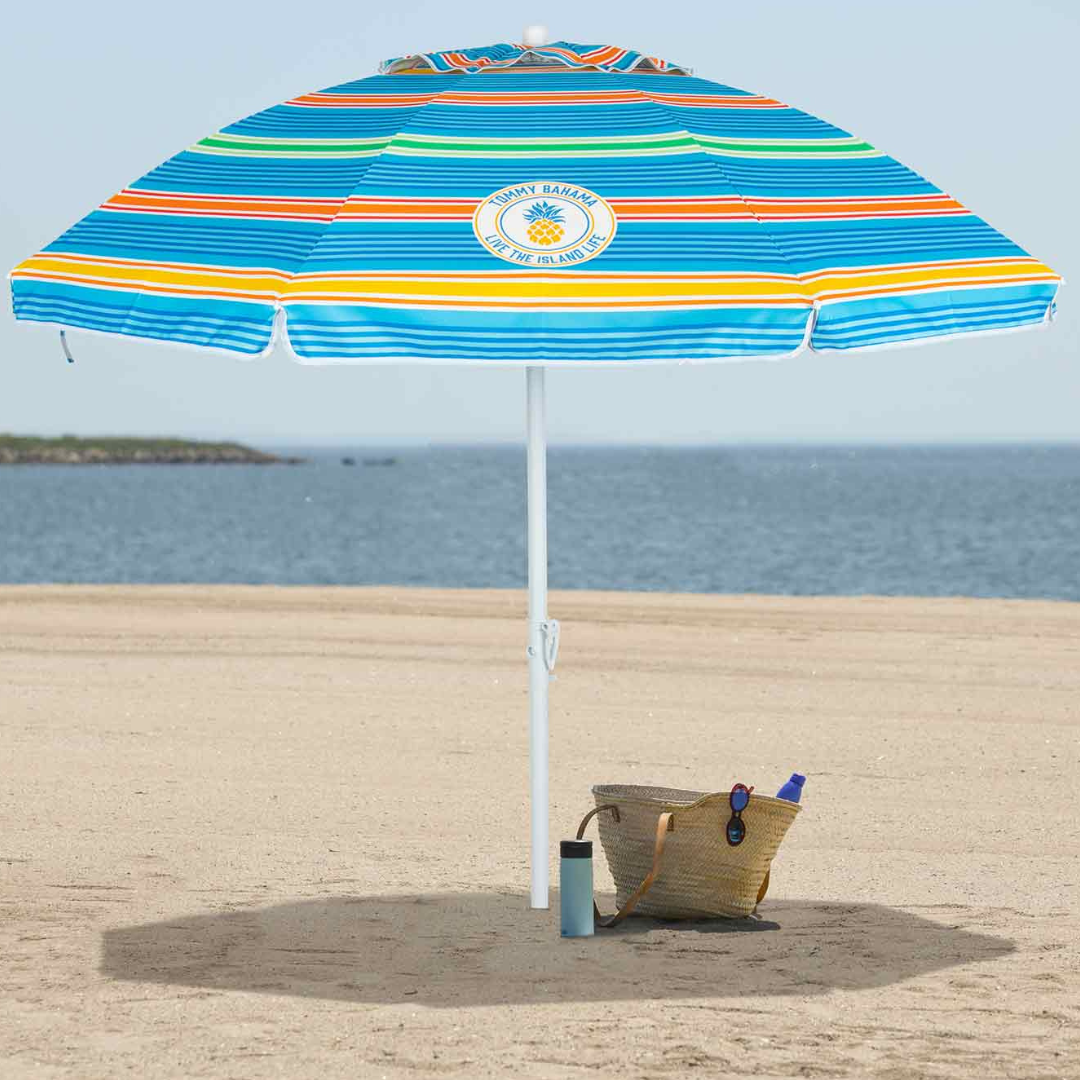 A colorful striped beach umbrella with the text \“Tommy Bahama, Live the Island Life\“ is set up on a sandy beach. Nearby is a woven straw bag containing a blue bottle and sunglasses, along with a tumbler on the sand.