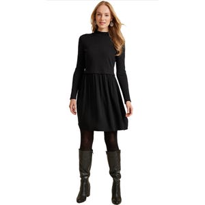 A woman in a black long-sleeve mini dress with a high collar. The dress has a fitted top and a slightly flared skirt. She is wearing black tights and knee-high black boots.