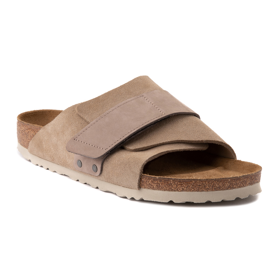 A pair of beige suede slide sandals with an adjustable strap and cork sole.