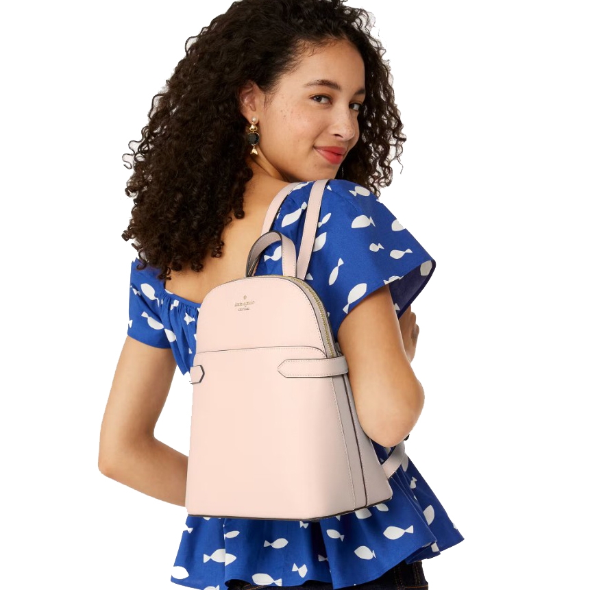 A woman is wearing a blue top with white patterns and carrying a light pink Kate Spade backpack on her shoulder.