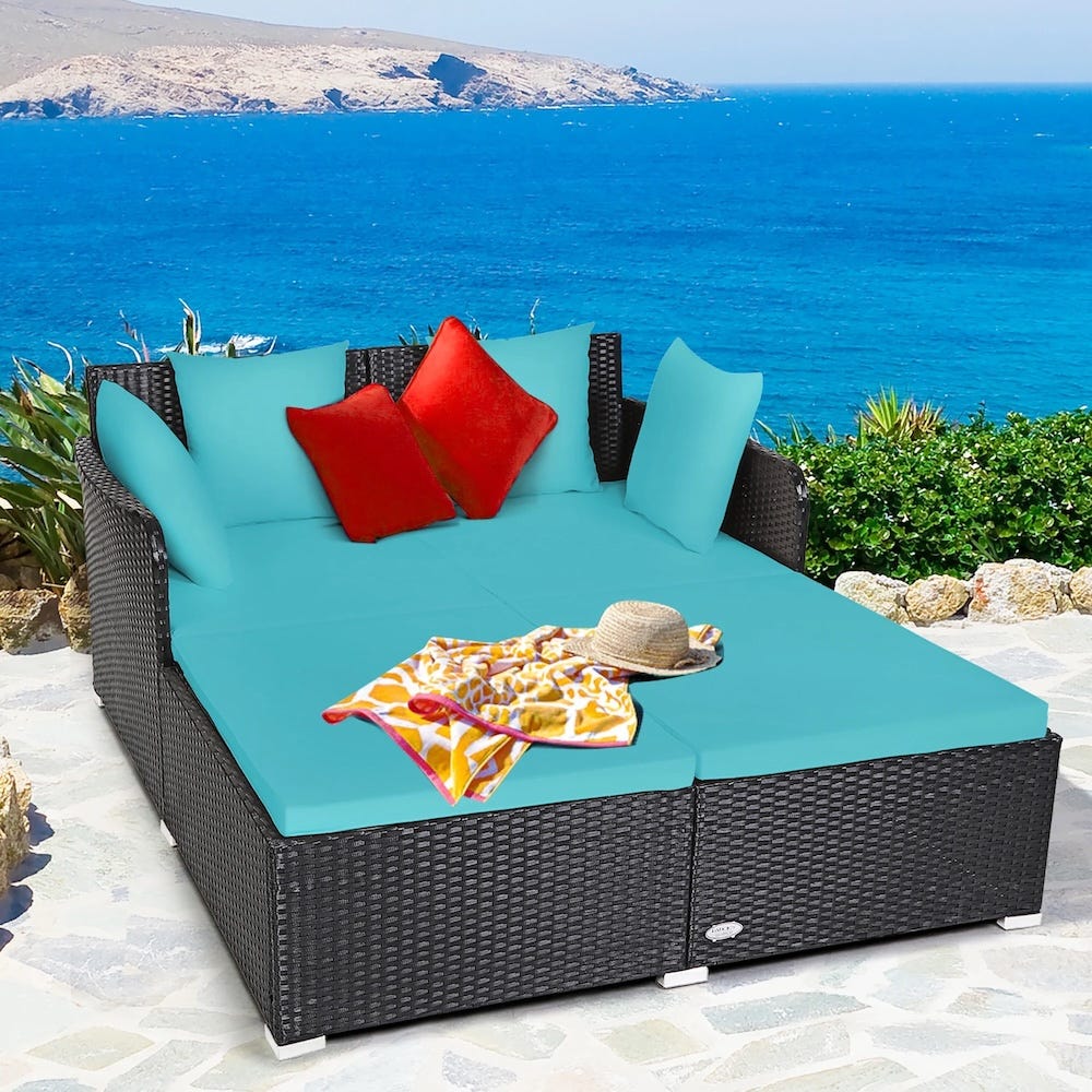 Outdoor wicker daybed with turquoise cushions, red accent pillows, and a straw hat and colorful towel on top, set against a seaside backdrop.