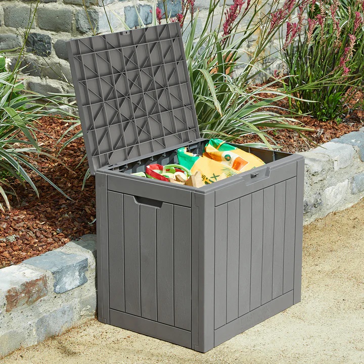 A gray plastic storage box with its lid open, showing colorful outdoor toys inside, set in a garden environment.