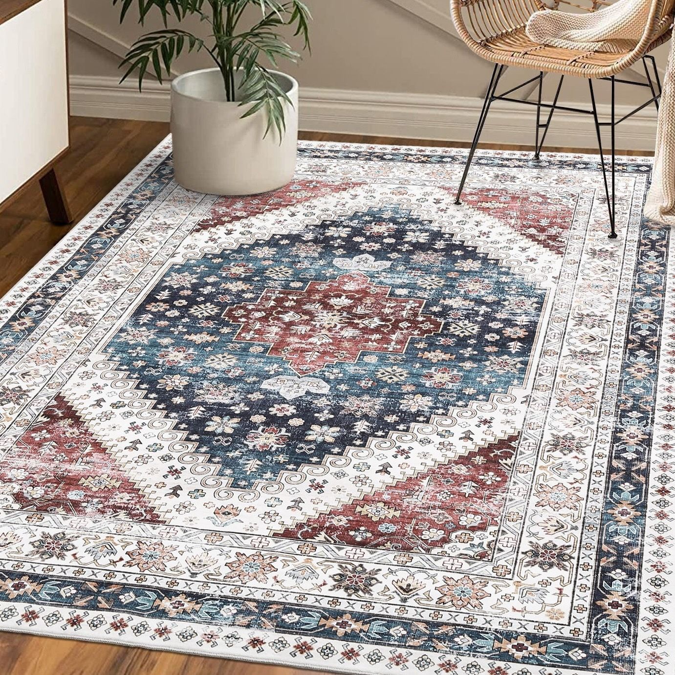 A rectangular area rug with a traditional pattern features blue, red, and white colors. It's placed on a wooden floor, accompanied by a potted plant and a woven chair.