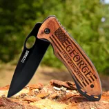A folding pocket knife with a black blade and wooden handle engraved with \“George\“ and \“Love You Pops,\“ along with an American flag design.