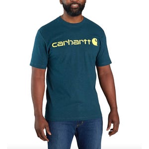 A man is wearing a teal Carhartt T-shirt with yellow logo text across the chest, paired with blue jeans.