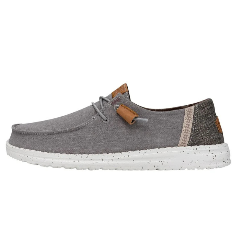 A pair of Hey Dude Wendy Washed Shoes featuring a grey canvas upper with a white sole and leather-look tab on the heel. The shoe has lace-up detailing and a patterned band accent.