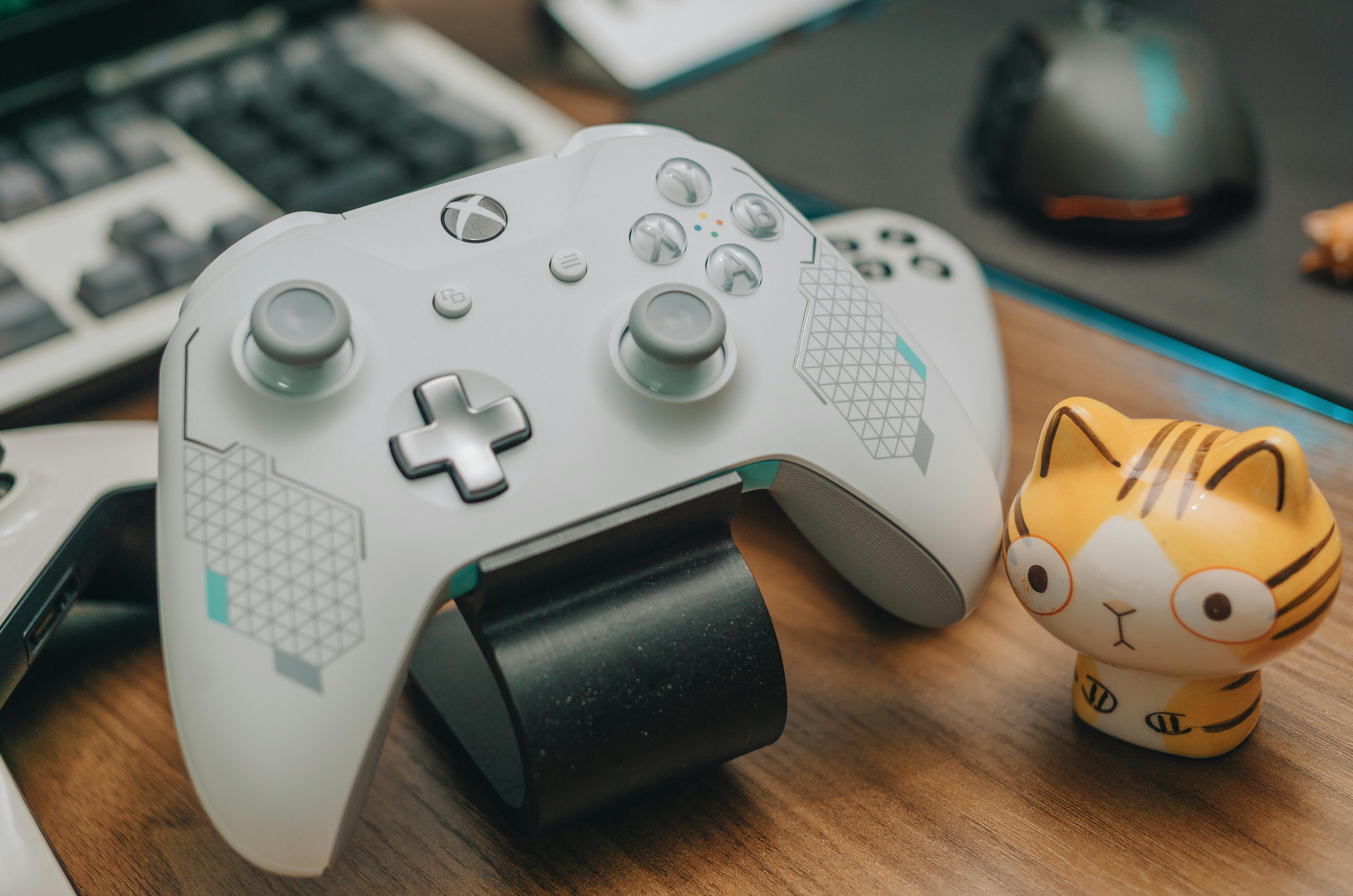 A white Xbox controller, a computer mouse, and a small figurine of a striped cat on a desk.