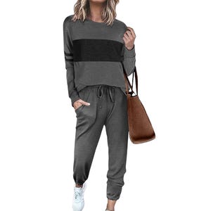 A woman is wearing a casual two-piece loungewear set featuring a long-sleeve top with a black stripe across the chest and black athletic stripe details on the sleeve, paired with matching drawstring pants and white sneakers. She's holding a brown tote bag.