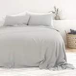 A bed with light gray sheets and pillows is set against a white wall. A striped basket with cushions is placed on a rug beside the bed, along with decorative vases and a small plant on the right.
