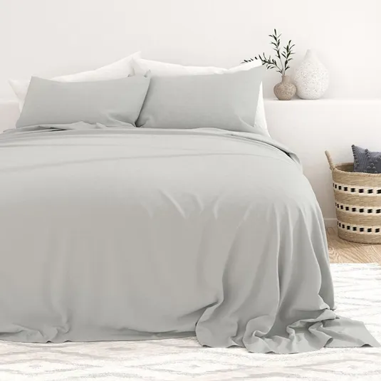 A bed with light gray sheets and pillows is set against a white wall. A striped basket with cushions is placed on a rug beside the bed, along with decorative vases and a small plant on the right.