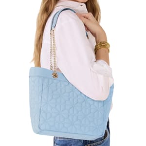 Woman holding a light blue quilted tote bag with a chain strap, wearing a white blouse and blue jeans with gold bracelets.