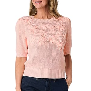 Light pink knit floral sweater top with short sleeves, features a floral pattern on the chest. Ribbed hem and sleeves add a classic touch. Worn with dark jeans.