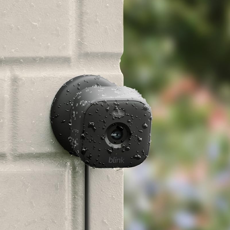 A wall-mounted, black, outdoor security camera with rain droplets on it.