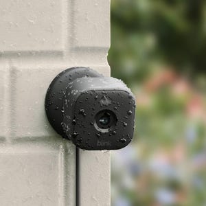 A wall-mounted, black, outdoor security camera with rain droplets on it.