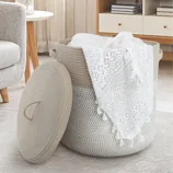 A woven rope storage basket with a lid, holding a white lace cloth, is set in a cozy living room with a sofa, round table, and a bookshelf in the background.