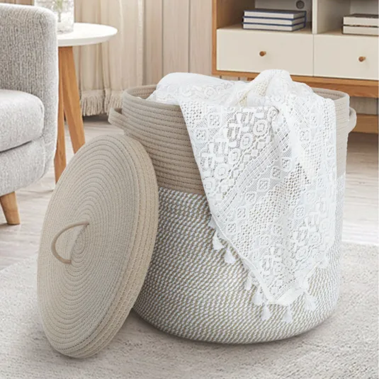 A woven rope storage basket with a lid, holding a white lace cloth, is set in a cozy living room with a sofa, round table, and a bookshelf in the background.
