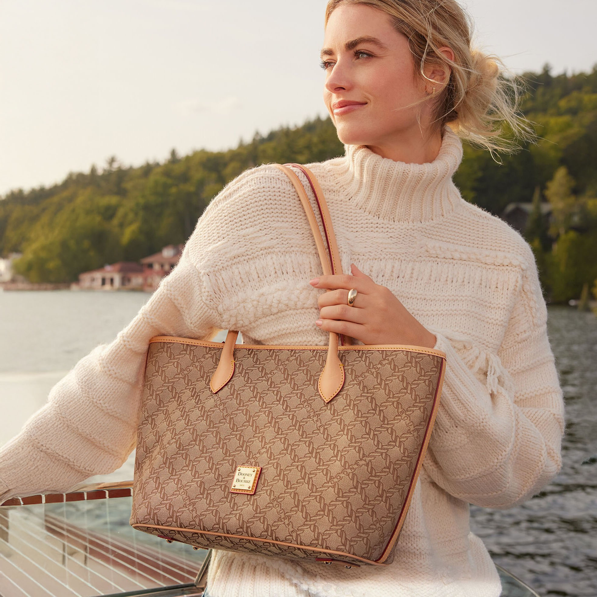 The Dooney & Bourke Maritime Tote features a beige monogram pattern with tan leather accents and straps, and includes a small gold logo patch on the front. The woman carries the tote while wearing a cozy cream turtleneck sweater by a serene lakeside setting.